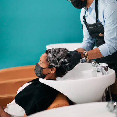 Male hairstylist washing African American woman's hair at reopened salon during coronavirus pandemic.